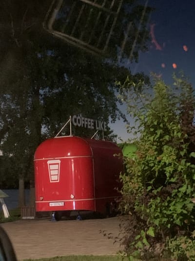The image shows a red coffee truck parked on a paved surface with some grass and foliage surrounding it. The truck has a white logo of a coffee cup on its side. Above the truck, a sign reads "COFFEE LIKE". In the background, there are trees and the night sky with some lights visible. A license plate on the truck is partially visible and appears to read "BT 0576". The overall scene suggests an outdoor food vendor operation, possibly in a park or public area. No people are visible in the image. The location in the city of Krasny Klyuch, Russia is mentioned, but there are no visual cues in the image to confirm or deny this specific location.