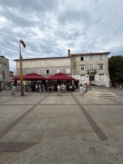 In the city of Krk, Croatia, an overcast sky looms over a paved town square. The square is bordered by a two-story building with a tan facade and white shutters, featuring multiple windows and a balcony on the second floor. To the left of this building, another stone building with similar windows stands. A tall wooden flagpole with a red and gold flag stands in the foreground. Underneath two large, red awnings, people are gathered at tables, likely at an outdoor cafe or restaurant. A stone well with intricate carvings is situated in the center of the square. Several people are walking around the square, some interacting with each other, while others are seated at the outdoor tables. The ground is paved with large square tiles, with red rectangular tiles forming a pattern. The scene depicts a typical day in a European town square, with people enjoying the outdoors amidst historical architecture.