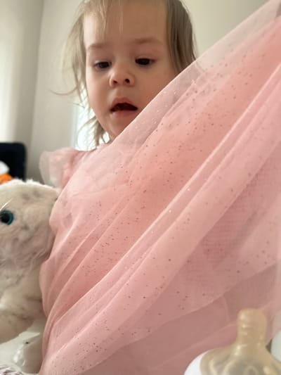 A child with light hair is peering out from behind a pink, glittery tulle fabric. The child has an open mouth and is looking downwards. A white plush toy is visible beneath the fabric. A baby bottle is visible on the right. The scene appears to be indoors. The location in the city of Kharnavy, Russia, is unknown.