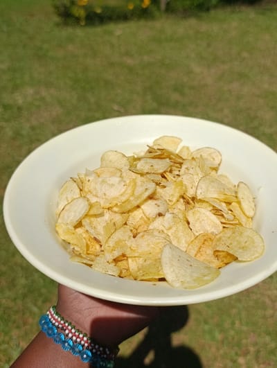 A close-up image shows a person's hand holding a bowl filled with potato chips. The hand is dark-skinned and is adorned with two distinct beaded bracelets: one features horizontal stripes of green, white, and red beads, while the other is composed of larger, translucent light blue beads. The bowl itself is a simple, light-colored (cream or pale yellow) dish, brimming with golden-yellow, thinly sliced potato crisps.
The setting is outdoors, under bright daylight. The background is dominated by green grass, which appears lush and well-maintained, suggesting a garden, lawn, or park environment in Kaplelwo, Kenya. In the far background, out of focus, some small yellow or orange flowers can be vaguely seen among the greenery. The strong sunlight casts a distinct shadow of the hand and bowl onto the grass, indicating a sunny day, likely during the midday or early afternoon hours. No specific activity beyond holding the bowl of chips is depicted, and no text is visible in the frame.