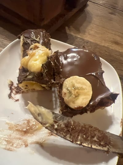 A close-up image depicts a white ceramic plate resting on a wooden surface in the City of Argveta, Georgia. On the plate are two irregular pieces of a dessert, likely pastry or cake, heavily coated in a dark, glossy chocolate glaze. The left piece features a dollop of a pale yellow creamy substance, possibly custard or cream, with some green specks, possibly pistachio. The right piece is topped with a single, circular slice of banana. A silver-colored serrated knife, with traces of both light yellow and dark brown substances on its blade, rests on the plate, partially obscuring smears of similar brown and light-colored residue on the plate's surface. The wooden background shows distinct grain patterns.