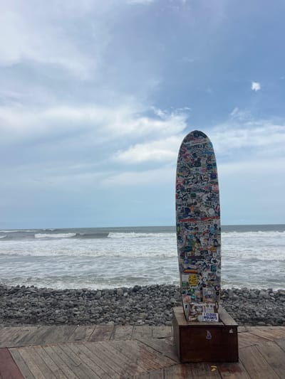 The image depicts a surfboard covered in numerous stickers, standing upright on a wooden platform near a rocky beach. The ocean with waves is visible in the background under a cloudy sky. The surfboard is positioned on top of a wooden crate. A sign on the crate reads "I CLOSED WOLSKI'S MILW, WISC." The scene is located in El Tunco, El Salvador.
