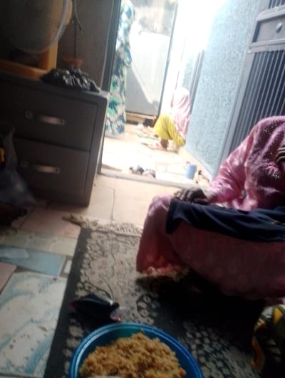 The scene depicts an indoor setting, possibly a residence, in the city of Dorayi, Nigeria, transitioning to an outdoor area visible through an open doorway. In the foreground, a blue plate containing jollof rice and what appears to be stewed chicken is positioned on a patterned rug. Partially visible, an individual in a pink garment with a dark blue fabric draped over their lap is seated or reclining on the rug, to the right of the food plate. Their hands are visible on the blue fabric. To the left of the food, a small black object resembling a pouch or bottle lies on the rug.
To the left of the foreground, a dark two-drawer metal cabinet is visible. Above it, a portion of a brown object, possibly a fan or a shelf, is present. Further back, near an open doorway leading outdoors, two more individuals are visible. One individual, dressed in a white and green patterned gown, stands near the doorway frame. Another individual, wearing yellow trousers and a light pink head covering, is seen stooping or kneeling on the tiled floor just outside the doorway. A blue cup is on the floor near this individual. The outdoor area beyond the doorway features a concrete or tiled surface and what appears to be a metal gate or wall structure. The lighting suggests a mix of indoor ambient light and brighter light from the outdoor area. The floor inside is a combination of light-colored patterned tiles and the aforementioned rug.