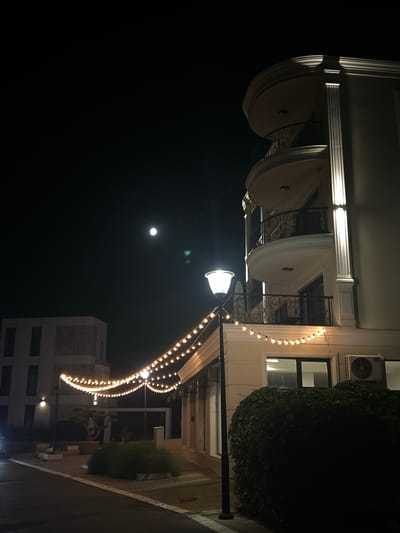 An outdoor night scene in Chernomorets, Bulgaria, features a modern, multi-story building with decorative columns and curved balconies, illuminated by subtle exterior lighting. A string of warm-toned lights drapes across the foreground, connecting two lampposts. In the distance, another building with dark rectangular windows is visible under a dark sky with a full moon. Lush green bushes and planters are present in the foreground, adjacent to a paved pathway and street. An air conditioning unit is mounted on the exterior wall of the main building. The overall impression is one of a tranquil, illuminated night setting.