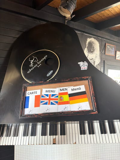 This is a medium-angle, eye-level, full shot of a grand piano with a black finish, situated in what appears to be an indoor establishment. The piano's keys are visible in the foreground, with some showing signs of wear and tear. Mounted on the piano's lid is a framed display containing several cards, each featuring a flag and text. From left to right, the visible flags are those of France, the United Kingdom, Spain, and Germany, with corresponding text labels such as "CARTE," "MENU," and "Menü." A circular black drumhead with white lettering and a logo that reads "doobop BAR" is also attached to the piano's lid, slightly above and to the left of the framed cards. In the background, a black wooden wall paneling is visible, and a portion of a ceiling with wooden planks and a spotlight fixture is also in view. To the right of the piano, a doorway or window reveals a bright outdoor area. A large black and white poster featuring a stylized portrait of a person is affixed to the wall. The establishment is confirmed to be "Doo Bop Bar" in Espinho, Portugal, based on the information provided. There are no people visible in the image.
