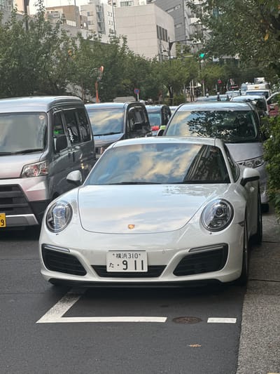 A white Porsche 911 coupe is parked on an urban street in Chūō, Japan, oriented facing forward. The vehicle's front displays a Japanese license plate reading "横浜310 た ・9 11", indicating Yokohama registration. White L-shaped road markings define a parking space directly in front of the Porsche on the dark asphalt pavement. A circular metal manhole cover is present on the asphalt to the right of the parking markings.

To the immediate left of the Porsche, a silver Japanese kei van or minivan is parked, partially obscuring its yellow license plate. A line of additional vehicles, including other minivans and what appear to be sedans or SUVs, extends down the street behind the Porsche, suggesting either queued traffic or parallel parking.

The environment includes green trees and bushes lining the right side of the street and along the sidewalk, with some foliage reflecting in the vehicles' surfaces. Multi-story, light-colored urban buildings with numerous windows form the background. Street infrastructure visible includes a red and white striped pole with a convex mirror and a green traffic signal in the distance. No discernible people are present in the foreground. The sky, reflected in the car's windshield and body, appears overcast.