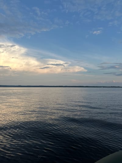 The image depicts a wide expanse of calm water under a partly cloudy sky. The water is dark blue and reflects the muted colors of the sky, with gentle ripples across its surface. In the distance, a dark, low-lying horizon is visible, suggesting land with a tree line. The sky is a blend of light blue, white, and pale yellow clouds, with some streaks of cirrus clouds. The lighting suggests it may be late afternoon or early evening, with the sun casting a soft glow. The image appears to be taken from a boat or a low vantage point near the water. There are no people or specific man-made structures clearly visible, except for a faint white object that could be a small boat or a building in the far distance on the horizon. The overall atmosphere is serene and expansive. The location is specified as Nozhovka, Russia, implying this scene is set within or near that geographical area.