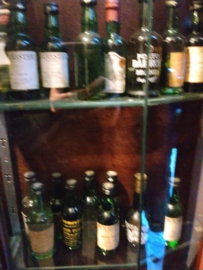 A glass cabinet displays an assortment of vintage liquor bottles on two shelves. The upper shelf features bottles with prominent labels, some partially obscured. The lower shelf holds a collection of smaller, green bottles with aged labels, alongside bottles of varying colors, including one with a gold cap. The backdrop is a dark, wooden surface. The overall image has a slightly blurred, aged quality.