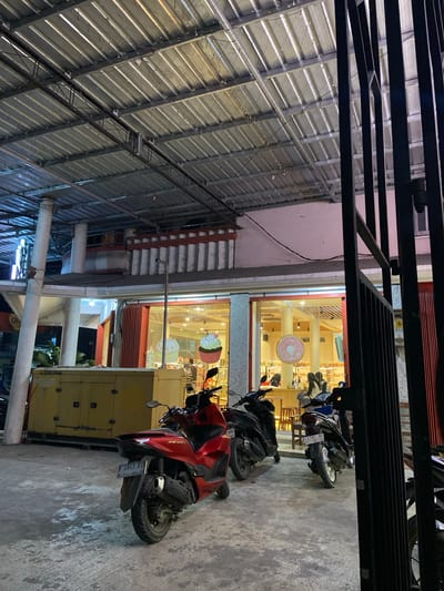 The image depicts the exterior of a bakery at night, with motorcycles parked in front. The bakery has a large glass storefront displaying baked goods and customers inside. A large yellow generator is parked to the left of the entrance. The bakery's signage is not clearly visible. The scene appears to be on a street in Crewek, Indonesia.