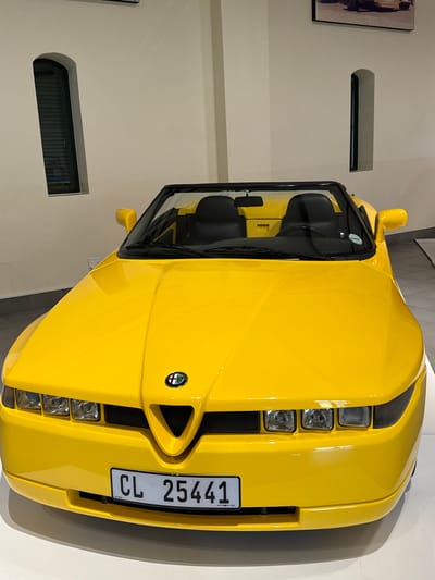 The image shows a yellow Alfa Romeo Spider parked indoors, presumably in a showroom or a private collection. The car is positioned at an angle, showcasing its front design. The license plate reads "CL 25441." The backdrop is a beige-colored wall with two recessed rectangular windows. A framed artwork is visible on the wall to the left. The location is indicated to be in Stellenbosch Local Municipality, South Africa.