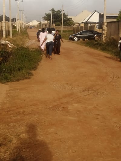 In Katon Rikkos, Nigeria, a group of five individuals is walking down a dirt road. Two women in the foreground are walking away from the camera. The woman on the left is wearing a white top and blue pants, while the woman on the right is wearing a dark dress. Behind them, three other individuals are walking towards the camera. One is wearing a light pink garment, another is wearing a black motorcycle helmet and a dark outfit, and the third is dressed in white. A black sedan is parked on the right side of the road, partially obscured by overgrown grass and a stone wall. The road is flanked by sparse vegetation and concrete utility poles with wires. Houses with distinctive rooflines are visible in the background, suggesting a residential area within the city. The sky is overcast, indicating a potentially humid or rainy day.