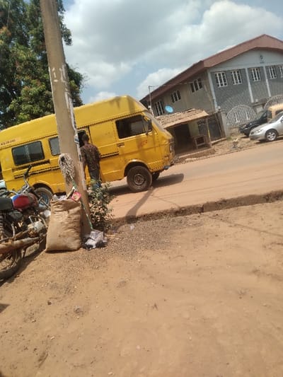 This is a daytime street scene in Agege, Nigeria, under a partly cloudy sky.

In the left foreground, a red and black motorcycle is partially visible, parked on a reddish-brown dirt and gravel surface. Adjacent to it stands a light-colored utility pole with white markings. Leaning against shrubbery near the pole is a large, light-brown sack.

A prominent yellow commercial van, a type of minibus, is positioned on a paved road that traverses the mid-ground from left to right. The van faces right and is slightly angled away from the viewer. A male individual, wearing a patterned shirt and blue trousers, stands next to the driver's side of the yellow van, facing away from the camera. The paved road features a raised curb separating it from the unpaved shoulder.

In the background, a multi-story building with a reddish-brown tiled roof and a grey facade dominates the right side. It features multiple white-framed windows and a distinctive white crest or logo near the roofline. A blue satellite dish is mounted on a pole near this building. Several parked vehicles, including a dark sedan and a silver sedan, are visible on the road or adjacent parking area near the main building. A smaller, darker-roofed structure is also visible behind the yellow van.

The immediate ground in the foreground and the road shoulder consists of reddish-brown dirt, gravel, and scattered debris, transitioning to a paved road surface.