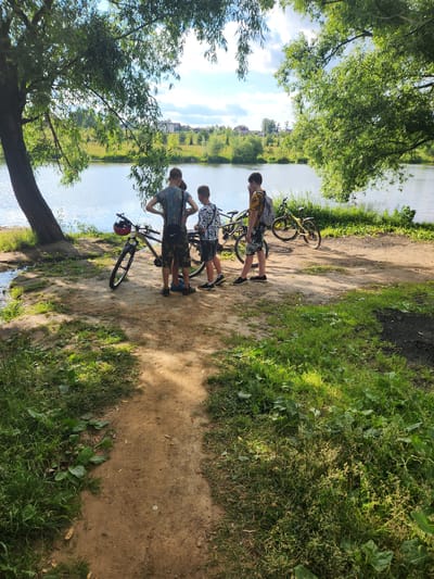 Three young males, appearing to be adolescents, stand near three bicycles by the edge of a tranquil body of water, possibly a lake or river, in Vniissok, Russia. The scene is set under the dappled shade of large, leafy trees. The ground is a mixture of sand and dirt, with patches of green grass and vegetation. In the background, across the water, a suburban landscape with houses and trees is visible. The sky is bright with scattered clouds, indicating a sunny day. One of the males is wearing a t-shirt with a camouflage pattern and dark shorts, while another wears a patterned t-shirt and dark shorts. The third male is seen from behind, wearing a t-shirt and dark shorts. One of the bicycles has a red helmet resting on its handlebars. The overall atmosphere suggests a casual outdoor activity, likely a bike ride near a natural setting within or on the outskirts of Vniissok.