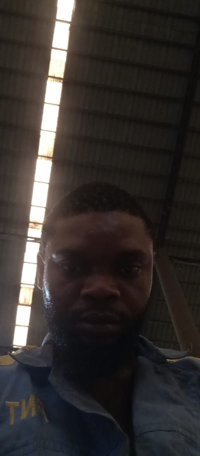 A close-up photo captures a dark-skinned male with a beard, wearing a blue uniform shirt with yellow embroidered letters. The background reveals a corrugated metal ceiling with a line of bright, rectangular skylights. The environment is dark except for the light streaming through the skylights, suggesting an interior space, possibly an industrial or commercial building. The image was taken in Magboro, Nigeria.