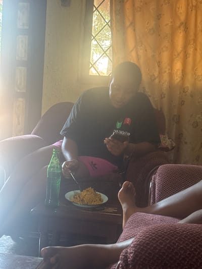 The scene depicts an interior setting, identified as a domestic residence in Aguata, Nigeria. A seated individual, partially silhouetted by bright sunlight entering from the left, occupies an armchair upholstered with a dark patterned fabric. This person wears a black t-shirt featuring a white "OFF-WHITE" logo and pink shorts with an Adidas emblem. They are simultaneously holding a smartphone in their left hand and a spoon in their right hand, which is positioned over a white bowl of a rice-based dish resting on a small wooden table. A green glass bottle also sits on this table. In the immediate foreground, the bare feet and lower legs of a second individual are visible, resting on another armchair with matching upholstery. A gridded window and floral-patterned curtains are discernible in the background. The room is strongly illuminated from the left, causing significant lens flare.