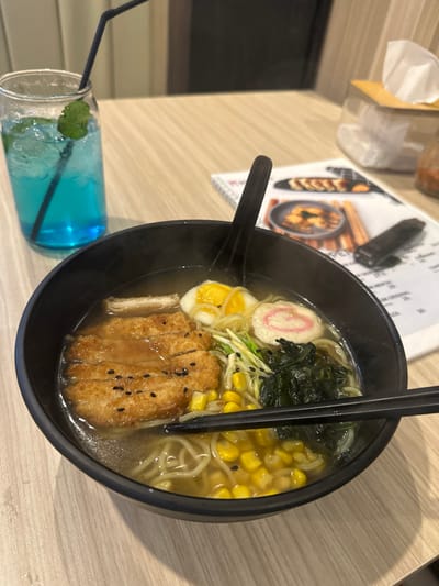 The image captures a close-up, overhead view of a meal served on a light wooden table, likely in a restaurant or cafe. The central focus is a steaming black bowl of ramen, presented slightly off-center to the right.
The ramen is rich with various ingredients: light-colored noodles submerged in a savory broth, topped prominently with a large piece of crispy, golden-brown katsu (fried cutlet) garnished with black sesame seeds. Beside the katsu is a halved hard-boiled egg with a vibrant yellow yolk. Other toppings include bright yellow corn kernels, dark green seaweed (possibly wakame), thinly sliced green onions or scallions, and a slice of narutomaki (a white fish cake with a distinctive pink swirl pattern). A black spoon rests in the bowl, while black chopsticks are placed across the rim. Visible steam rising from the bowl suggests the ramen is hot and freshly prepared.
To the lower left of the ramen bowl, a tall glass holds a vibrant blue beverage filled with ice cubes and a few mint leaves, with a black straw inserted.
In the background, on the same wooden table, a partially visible paper menu displays images of other dishes, including what appears to be dumplings or gyoza in a pan. A dark, rectangular object, possibly a wallet or phone, is resting on the menu. Further in the upper left, a portion of a light-colored tissue dispenser or box is visible. The lighting suggests an indoor setting, likely artificial. No people are visible in the frame, and any text on the menu is illegible. The image is captured at an angle, making the table appear tilted.
