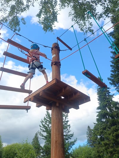 A young girl wearing a blue helmet, pink jacket, and grey shorts is navigating an obstacle course. She stands on a wooden platform, with one foot on a wooden beam that is part of a suspended bridge. The course consists of wooden planks and ropes, supported by a central wooden pole. The background shows a bright blue sky with scattered white clouds, and the tops of green trees. The image appears to be taken from a low angle, looking up at the girl and the course.