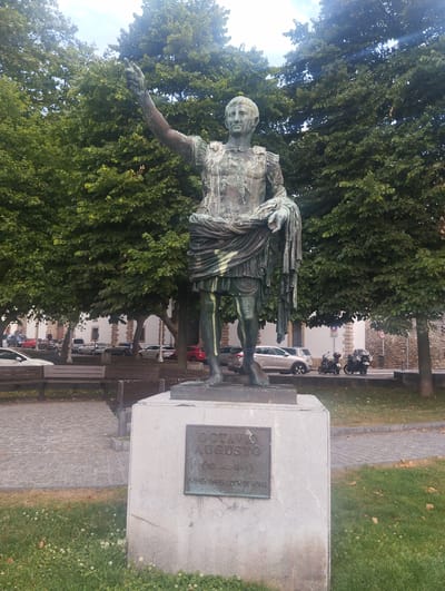 A bronze statue of a man in Roman military attire stands on a light gray stone pedestal. The statue depicts the figure with his right arm raised and pointing forward, and his left hand holding a draped cloth over his left arm. The statue is weathered and appears green with age. The pedestal has a metal plaque inscribed with text. The statue is situated in an outdoor urban environment with mature trees with green foliage providing a backdrop. In the background, there are several cars, motorcycles, and benches. A paved walkway and grassy areas are visible in the foreground. The statue is located in the city of Gijón, Spain.