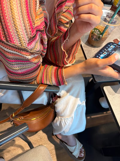 A top-down, eye-level shot shows a person seated at a table with a Starbucks iced drink and using a smartphone. The person is wearing a multicolored, striped knit sweater, and white pants. A brown leather purse is slung over the back of the chair. The person is wearing sandals on their feet. The location is a cafe in Spata-Loutsa Municipal Unit, Greece.