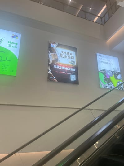The image shows a section of a shopping mall with escalators and illuminated advertisements on the wall. The advertisements are for a movie theater and a city youth center. One advertisement features a film strip and the text "大片驾到, 影有尽有 IMAX影城" which translates to "Blockbusters are coming, there are endless movies, IMAX Cinema". Another advertisement displays "城市年权力中心, 灵感漫游新聚场" which translates to "City Power Center, Inspirational Roaming New Gathering Place". The scene is indoors, likely a public space given the presence of escalators and the mall-like environment. The lighting suggests it is either daytime or the mall is well-lit internally. The location is identified as Liandu District, China. There are no people clearly visible in the frame. The focus is on the architecture and advertising within the mall.