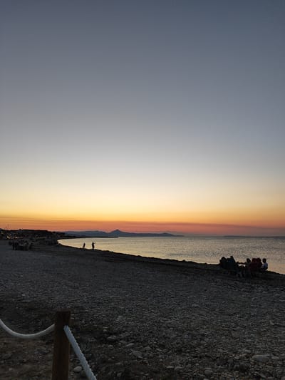 This is a landscape photograph taken at sunset on the pebble beach of Els Poblets, Spain. The sky transitions from a pale blue at the top to shades of orange and yellow near the horizon, indicating the setting sun. The sea is calm with gentle ripples. In the distance, a mountainous silhouette is visible against the colorful sky.
On the beach, several groups of people are present. In the mid-ground, to the left, there is a cluster of chairs and tables, suggesting a dining area or seating for a beach bar. Further down the beach, several individuals are visible, including two silhouettes standing near the water's edge. In the right mid-ground, a group of four people are seated in camping chairs, facing the sea. The foreground of the image shows a wooden post with a white rope tied to it, part of a boundary or barrier, and a textured surface of pebbles and stones that make up the beach. The lighting suggests it is twilight, with shadows lengthening and the overall scene illuminated by the fading sunlight.