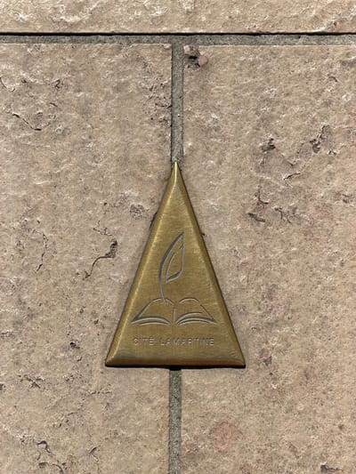 The image shows a close-up, top-down view of a brass triangular marker embedded in a tiled sidewalk. The marker is positioned in the center of the frame, with its apex pointing upwards. The brass has a warm, golden hue and is etched with a symbol that appears to be an open book with a leaf emerging from its pages. Below the symbol, the text "CITE LAMARTINE" is engraved. The sidewalk tiles are a pale, beige-brown color with a textured, stone-like appearance, and they are separated by dark grout lines. The lighting suggests it is daytime, casting subtle shadows. This marker is located on the Place Lamartine in Charnay-lès-Mâcon, France, a location significant to the poet Alphonse de Lamartine.