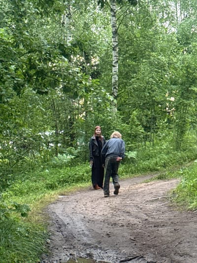 Two individuals are walking on a dirt path in a wooded area. The person on the left, with long reddish-brown hair, is wearing a dark, long coat and dark pants. The person on the right has lighter, curly hair and is wearing a dark gray jacket and dark pants. The path appears wet and muddy in places. The surroundings are densely vegetated with green trees and foliage, suggesting an outdoor, natural setting. The lighting indicates it is daytime. The location is identified as a wooded area within the city of Desnogorsk, Russia.