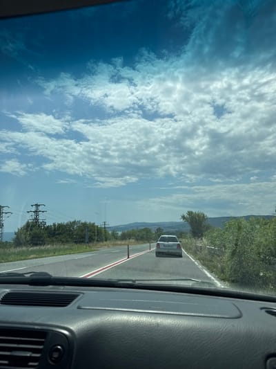 A forward-facing view from the interior of a car traveling on a road. The road has a white line and a red line painted on its surface. A white car with a license plate is visible ahead, also traveling on the same road. The landscape beside the road features green vegetation and trees, with power lines and towers in the distance. The sky is filled with scattered white clouds against a blue backdrop. The foreground shows the car's dashboard. The scene is consistent with a drive along the road leading towards the city of Byala, Bulgaria, potentially near the Black Sea coast given the distant water body.