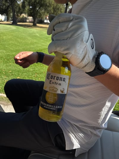 A person in a white golf glove and shirt holds a Corona Extra beer bottle at an angle. They wear a black wristwatch. The individual sits, possibly in a golf cart, wearing dark pants. A green lawn is visible in the background. The beer bottle's label is prominently displayed.