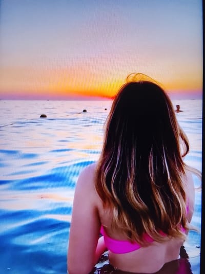 An over-the-shoulder shot shows a woman in a bright pink bikini from the waist up, facing a sunset over a body of water. The water is a vibrant blue with white and yellow highlights, suggesting ripples and reflections. In the distance, several buoys or markers are visible in the water, as well as a silhouetted person swimming or standing in the water. The sky transitions from a pale blue at the top to warm oranges, yellows, and pinks near the horizon, indicating either a sunrise or sunset. The woman's long, dark hair with lighter highlights cascades down her back. The scene appears to be a beach or coastal area. The image is cropped to focus on the woman and the sunset, with the background slightly out of focus.