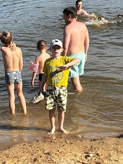 In Brilliant, Russia, a group of individuals are gathered at the edge of a body of water, likely a river or lake, on a sandy bank. The water is murky brown and appears calm with small ripples.
In the foreground, a young boy wearing a yellow t-shirt and camouflage shorts stands facing forward, holding a gray athletic shoe in his left hand. He has on a white baseball cap and glasses. To his left, another young boy in blue swim trunks with a moon and star pattern stands with his back to the camera. His left arm is resting on his shoulder. Behind him, a young girl with her hair in a bun is also facing away from the camera, wearing a dark swimsuit and a pink arm float with a floral pattern.
In the middle ground, a shirtless man with light-colored swim trunks stands facing the water. Further back, another shirtless man is partially submerged in the water, with another person swimming or playing in the water creating splashes. The background consists of more water and a sliver of land further away. The overall impression is a casual scene of people enjoying a warm day by the water.
