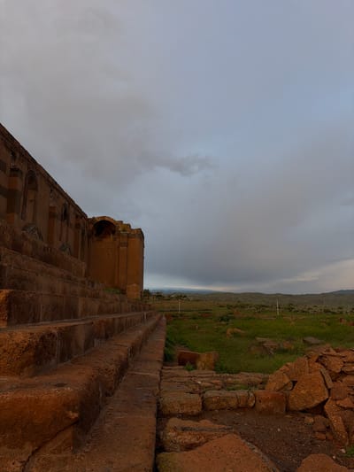 The image shows a partial view of a stone building, presumably a church or other religious structure, in Anipemza, Armenia. The structure has a curved archway, windows, and decorative patterns. Stone steps lead from the viewer towards the building. A landscape of green terrain and hills extends into the distance under a cloudy sky. A stone wall or path edges the green area.