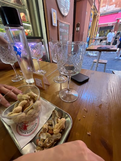 The image shows a table at a restaurant or bar in Bologna, Italy. The scene is captured from a high-angle perspective, looking down at the tabletop. Present on the table are several clear, ornate wine glasses, some empty and others possibly used. A small, rectangular black smartphone rests on the wooden surface of the table. A small paper menu card sits amongst the glasses. A silver tray contains a collection of peanuts, some still in their shells and others already opened. A clear plastic container also holds peanuts. A partially visible hand is present in the foreground of the scene. The environment suggests an outdoor seating area in the city, with pedestrians walking by and storefronts visible in the background.