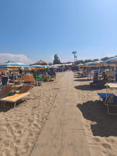 The image depicts a sunny day at a beach resort in San Salvo, Italy. A paved walkway runs through the sandy beach, lined with rows of beach chairs and umbrellas. Several people are visible relaxing on lounge chairs under the umbrellas, some appear to be sunbathing, while others are engaged in conversation. The sky is clear and blue with a few scattered clouds. In the background, there are trees and some structures that suggest the presence of amenities or facilities associated with the beach. The scene conveys a relaxed and leisurely atmosphere characteristic of a beach vacation.