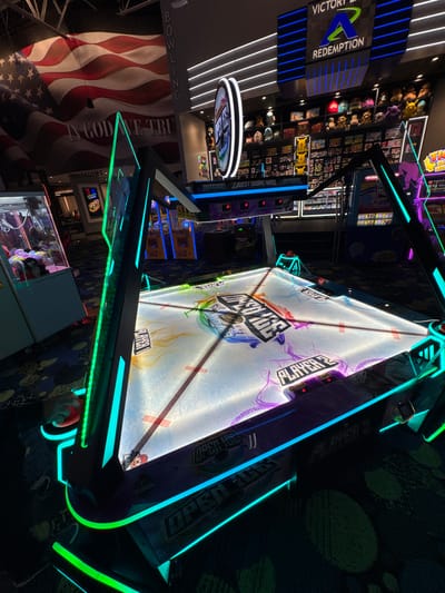 This image captures an indoor entertainment center, likely an arcade or a family entertainment complex in Schaumburg, United States of America. The scene is characterized by dim ambient lighting accentuated by vibrant neon lights from various arcade machines.
The central focus is a large, brightly lit multi-player air hockey-style game, possibly named "Open Ice" or "Open Air," featuring prominent blue and green LED strips along its edges. The playing surface is white with colorful graphics and displays showing "PLAYER 1," "PLAYER 2," "PLAYER 3," "PLAYER 4," "TIME," and "SCORE," all currently at '0'. A hand holding a red puck or controller is visible at the bottom right, suggesting active play or readiness to play.
To the left of the air hockey game, a sign for "VICTORY REDEMPTION" with a blue 'V' logo indicates a prize redemption center. Behind it, shelves are stocked with various plush toys and other arcade prizes, including a yellow Pikachu-like plush. Further left and below, a large American flag banner is displayed, vertically proclaiming "IN GOD WE TRUST" and "BOWLING," suggesting a bowling alley is part of the complex.
Other visible texts include "DEFENSE LOWEST SCORE WINS" on another game cabinet, and overhead signs for "ANDRECO DINING" and "RESTROOMS," confirming a multi-purpose entertainment venue. The overall atmosphere is lively and engaging, typical of an evening at an arcade.