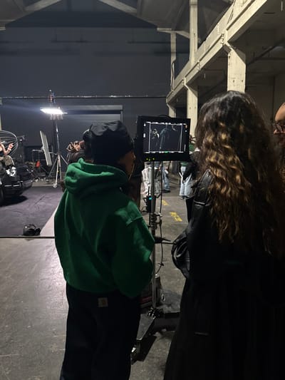 This image depicts an indoor film or photo production set located in Ivry-sur-Seine, France, characterized by a large industrial-style space with concrete floors, exposed concrete pillars and beams, and dark walls, indicative of a studio or warehouse conversion.

In the immediate foreground, facing right, is an individual wearing a black beanie, a green Carhartt hooded sweatshirt, and dark work pants with a visible Carhartt logo on the back pocket. This person is looking intently at a flat-screen monitor mounted on a stand, which displays a scene with two figures, one prominently in a dark coat or cape. To the right of the monitor, partially obscured, is another individual with long, wavy brown hair, wearing a dark jacket, also focused on the monitor. A third person, wearing glasses, is partially visible behind and to the right of the second individual.

The background reveals a bustling production environment. Various pieces of filmmaking equipment are visible, including large lighting fixtures on stands, a parabolic reflector, cables, and what appears to be a camera dolly or rig. Several indistinct figures are present in the deeper background, some standing and observing, one notably wearing a white protective suit. A large, dark roller door is visible against a dark wall in the mid-ground. The overall lighting suggests a controlled setup, with bright spotlights illuminating specific areas and the overall space appearing dim.