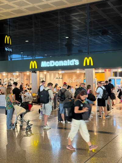 This is a slightly tilted, portrait-oriented photograph of a busy McDonald's and McCafe located in an airport terminal. The scene depicts a long queue of people waiting to order food. The McDonald's signage with its iconic golden arches and the "McCain's" logo is prominently displayed on the left side of the image. The interior of the restaurant is visible, with staff members serving customers and people sitting at tables in the background.
The subjects are diverse, including men, women, and possibly children, dressed in casual travel attire. Many are carrying backpacks and luggage, indicating they are either arriving or departing passengers. The overall atmosphere appears to be one of a typical busy travel hub, with a sense of anticipation and movement.
Notable details include the modern, sleek design of the airport terminal, with reflective floors and patterned walls. The lighting suggests it is daytime, possibly late afternoon, given the warm glow on the floor. There is no clear indication of weather conditions outside, but the indoor setting is well-lit and clean. The people in the queue appear patient and engaged, with no obvious signs of distress or frustration.
Visible text includes "McDonald's" and "McCafe" in their signature fonts. There are also some indistinct signs and menus within the restaurant, but the text is too small to be legible. The overall impression is a snapshot of everyday travel life, highlighting the universal presence of fast-food chains in major transportation hubs.