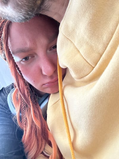 A close-up shot shows a person with reddish-orange braided hair and a yellow hoodie. The person is looking downwards with a sad expression. Part of a grey furry object, possibly an animal, is visible at the top of the frame. The person is wearing headphones around their neck. The scene appears to be indoors, possibly in a vehicle given the angle and lighting, but no specific city details can be discerned.