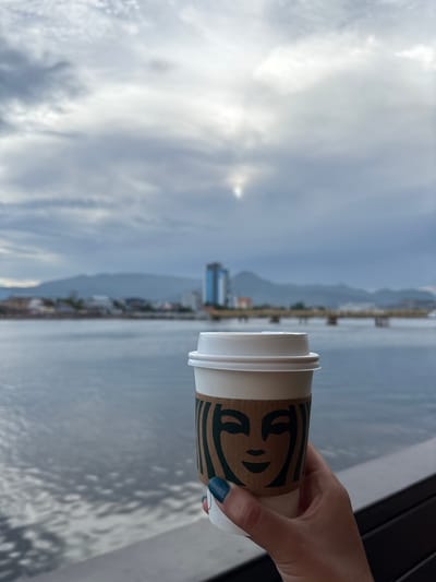 A hand with blue-speckled fingernail polish holds a Starbucks paper coffee cup with its signature green logo. The cup is positioned in the foreground, obscuring part of the view. Beyond the cup, a body of water stretches to the horizon, reflecting a cloudy sky. In the middle distance, a coastline is visible with buildings and a pier. Further in the background, mountains rise against the sky. The overall scene suggests a relaxed moment, possibly during daytime, with overcast weather. The presence of the Starbucks cup and the distant shoreline with buildings are the main identifiable elements in this media file.