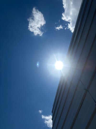 The image depicts a low-angle view of a bright sun shining against a clear blue sky. A portion of a modern building, possibly made of corrugated metal siding, occupies the right side of the frame, running diagonally from the lower right to the upper right corner. A lens flare from the sun is visible in the upper middle section of the sky. Several small, wispy white clouds are scattered throughout the sky, one prominent cloud cluster is near the upper left. The scene appears to be an outdoor environment within the Village of Johnson City, United States of America. There are no people visible in the image.