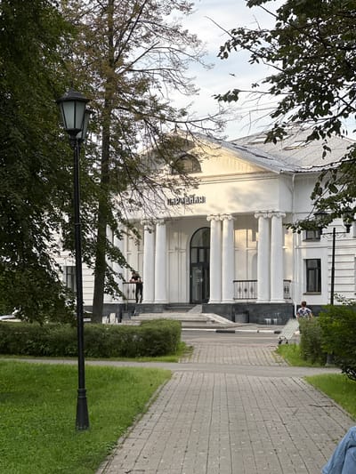 In Putyatino (Semivragovskaya S/A), Russia, a white building with a classical facade stands behind a paved walkway and manicured green bushes. The building features prominent white columns and an arched entrance. A black lamppost with an ornate design is positioned to the left of the walkway. Two people are visible: one individual stands near the building's entrance, leaning on a railing, and another person is seen pushing a wheeled cart on the right side of the walkway. Trees with green and yellowing leaves flank the scene, and the sky is visible above, with a bird in flight. The lighting suggests it is daytime, possibly late afternoon.