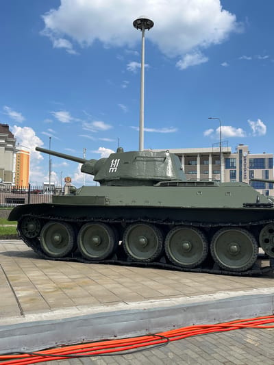 This image captures a T-34 tank, a Soviet-era medium tank, on display outdoors in Verkhnyaya Pyshma, Russia. The tank is painted olive drab and is positioned on a paved area, with its tracks visible. A white emblem with what appears to be Cyrillic characters is painted on the turret. In the background, several modern buildings are visible, including one with the word "KRATOVM" on its facade, suggesting an urban setting. The sky is blue with scattered white clouds, and a tall lamppost stands prominently near the tank. The foreground shows red corrugated tubes lying on the pavement. The scene appears to be a sunny day, possibly in a park or museum dedicated to military history.