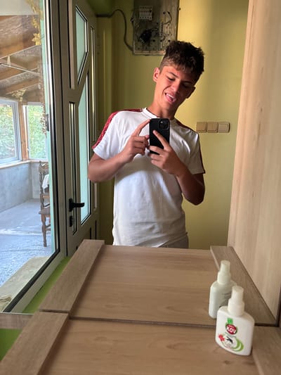 In Bulgarovo, Bulgaria, a young man is reflected in a mirror, taking a selfie with his phone. He is wearing a white t-shirt with red accents on the sleeves and is sticking his tongue out. The background shows a yellow wall with electrical outlets and a fuse box, a window with a view of greenery, and a wooden wardrobe. In the foreground, the mirror reflects two white bottles, one of which is labeled "KOV INSECTICIDE". The lighting suggests it is daytime.