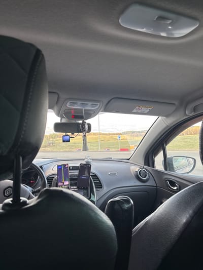 The image depicts the interior of a car, likely a Renault, as indicated by the steering wheel emblem. The view is from the back seat, looking towards the front. The dashboard is visible, along with two smartphones mounted on the air vents, displaying what appears to be navigation apps. A rearview mirror is positioned between the front seats with a dash camera attached to it. The car's interior is predominantly grey. The view through the windshield shows a landscape with green fields, a body of water, and some infrastructure, including parking signs and possibly buildings in the distance. Based on this and the fact that the model asked for Ermolaevo, Russia, the location of the photo is likely Ermolaevo, Russia.