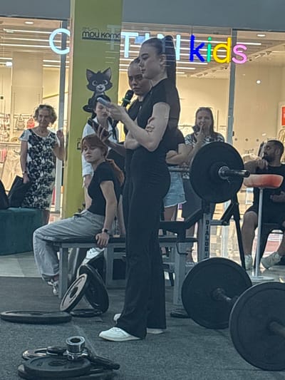 The image depicts an outdoor scene, likely in a public space. A group of people are gathered around weightlifting equipment. In the foreground, several weight plates are stacked on the floor, and a barbell is set up on a rack. Several individuals are visible, including a woman standing and looking at her phone, another woman sitting on a bench, and others in the background. Behind the group, a storefront with neon signage that reads "Kids" and "Tri Protoka" is visible. The overall environment appears to be an urban setting, possibly a shopping area or a community event.