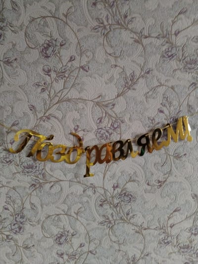 A golden banner spelling "Поздравляем!" (Congratulations!) in Cyrillic script is suspended across a wall with a floral embossed wallpaper. The wallpaper features a pattern of stylized flowers and scrolling leaves in muted tones of white, grey, and pale purple. The banner is made of a reflective, metallic material, likely plastic or acrylic, and is strung on a thin cord, partially visible. The banner's letters are connected in a cursive style. The overall impression is one of festive decoration. The image was taken in the city of Troitskoye, Russia.