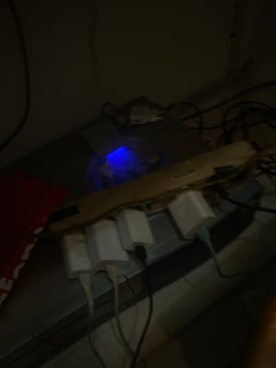 The image depicts a dimly lit indoor scene, specifically an area with electronic equipment, located in Cimenyan, Indonesia. No individuals are present in the frame.
In the center, a dark, rectangular electronic device, possibly a computer tower or game console, exhibits a prominent internal blue light, likely from a cooling fan or indicator. To the immediate right of this device, a beige or light-colored power strip is positioned horizontally, resting on a surface. Four white, rectangular power adapters/chargers are plugged into this power strip. Multiple cables extend from these adapters; at least two light green cables and one off-white cable are visible originating from the bottom of the chargers, leading downwards and to the right, connecting to unseen devices. Numerous dark cables are tangled behind and around the primary electronic device and power strip, indicating a cluttered setup.
On the far left, a portion of a red object, possibly fabric or a bag, is visible. The background consists of a plain, light-colored wall or surface. The overall environment appears to be a corner or desk area dedicated to electronic components and their associated power connections.