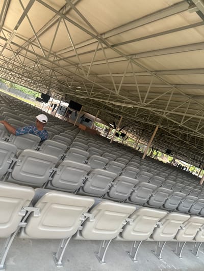 The image depicts a wide-angle view of a large, mostly empty, covered outdoor or semi-enclosed venue located in Ozuaha, Nigeria. Numerous rows of light beige or grey individual stadium-style seats, with flip-up bases, are arranged in a tiered formation, extending from the foreground into the background. Most seats are unoccupied, with their bases in the upright position.
In the immediate foreground on the left, a single male individual is seated in the first row, facing away from the camera towards the right side of the frame. He wears a white baseball cap and a patterned, short-sleeved shirt, with his right forearm resting on the top edge of the seat back to his immediate right.
Above the seating area, an expansive light-colored canopy roof is visible, supported by a dense network of grey metal trusswork. Fluorescent light fixtures are suspended from this structure. Towards the mid-ground, centered to the right, are structures resembling a stage or platform. Here, multiple large, rectangular black screens or monitors are mounted, flanked by audio speakers and additional supporting framework.
Beyond the structured seating and stage area, green trees and foliage are present, indicating an outdoor environment. A small, indistinct group of individuals is faintly visible in the far background on the right. The floor beneath the seats appears to be concrete or a similar hard, grey surface. The scene is illuminated by natural light, suggesting daytime.