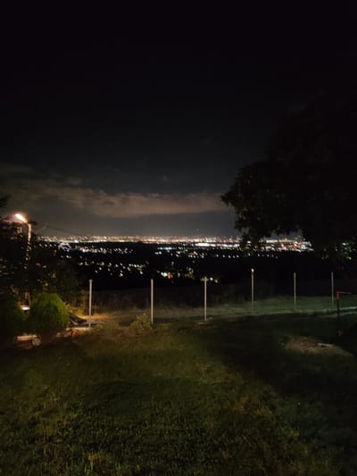 The image presents a nighttime panoramic view of the city of Dragovishtitsa, Bulgaria, from an elevated perspective. The upper portion of the frame shows a dark night sky with prominent, faintly illuminated clouds appearing in shades of gray and light brown, particularly towards the left and center. A continuous band of blurred and horizontally streaked artificial lights, characteristic of a sprawling urban area, stretches across the distant horizon, suggesting either motion blur or an out-of-focus capture.
In the immediate foreground, a dark green grassy area is visible, with patches illuminated by a yellowish-green light source. A chain-link fence, supported by several slender white vertical posts, runs horizontally across the lower mid-ground, marking the edge of the viewpoint. To the far left, a tall street lamp stands, emitting a bright, warm white light. Beneath and slightly to the right of the lamp, a small, rounded evergreen shrub or bush is partially visible and illuminated. The right side of the frame is dominated by the dense, dark foliage of a large tree, which extends from the top right corner down into the mid-ground, partially obscuring the distant city. A faint red object is discernible behind the fence towards the right edge. The scene is devoid of human figures or discernible activity.
