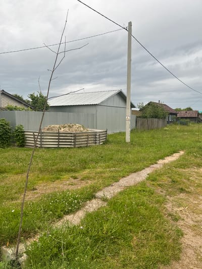 The image shows a cloudy day in a rural setting in Elmaachi, Russia. The scene features a path or dirt road running along a grassy area with a concrete border. In the background, there are several buildings, including a long, grey metal shed with a corrugated roof, and a more traditional wooden house with a reddish-brown roof. A wooden fence separates the properties in the distance. A concrete utility pole with electrical wires runs through the frame, and a bare-branched tree is visible in the foreground. There is a small, grey, corrugated structure holding a pile of what appears to be hay or straw. The overall impression is one of a quiet, somewhat rustic village. There is a yellow warning sign on the utility pole with black text and a symbol.